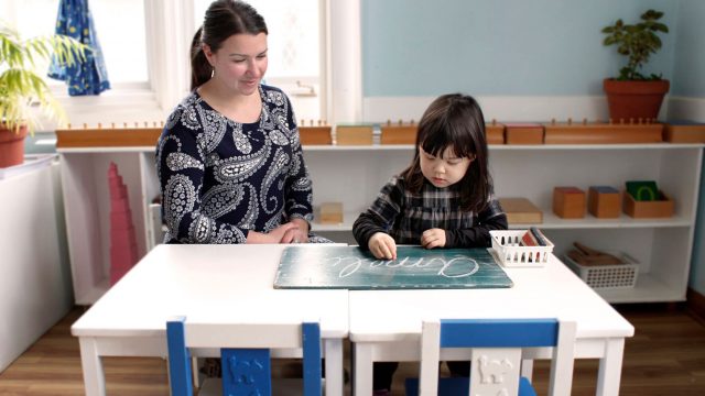 A teacher sitting with a little girl practicing her penmanship on a chalkboard