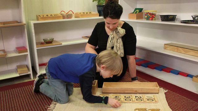 Teacher sitting on the floor with a little boy, using a wooden toy with numbers