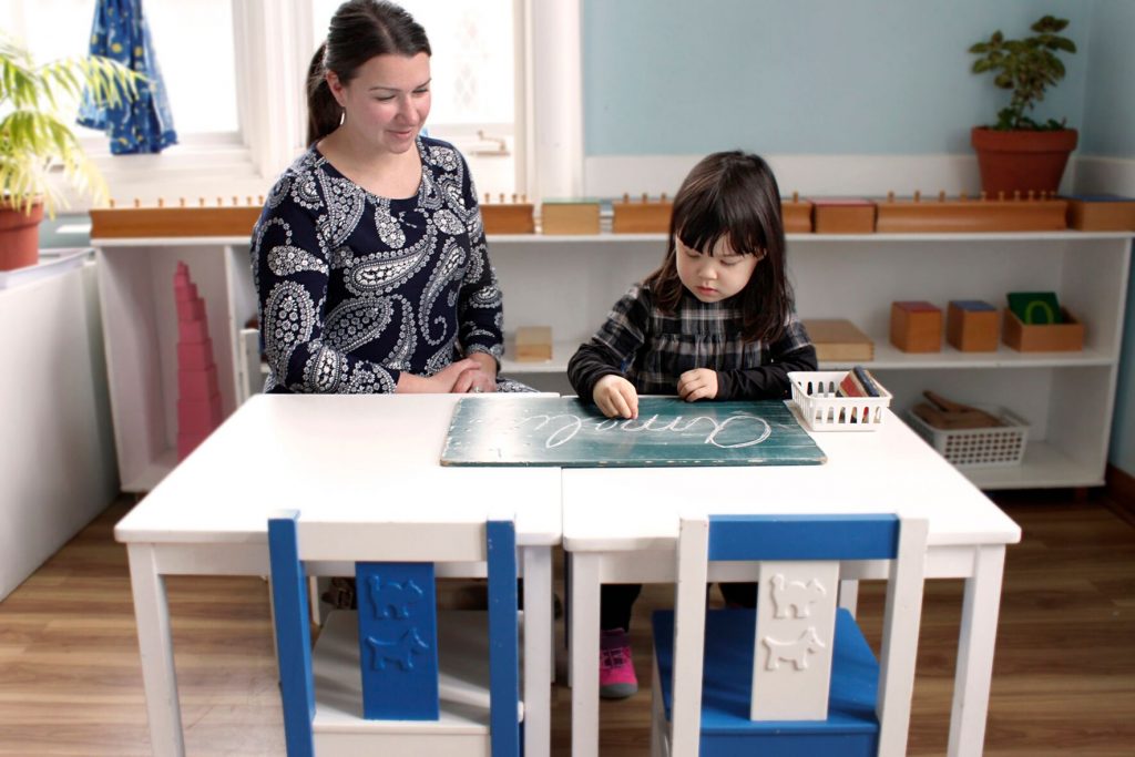 A teacher sitting with a little girl practicing her cursive on a chalkboard